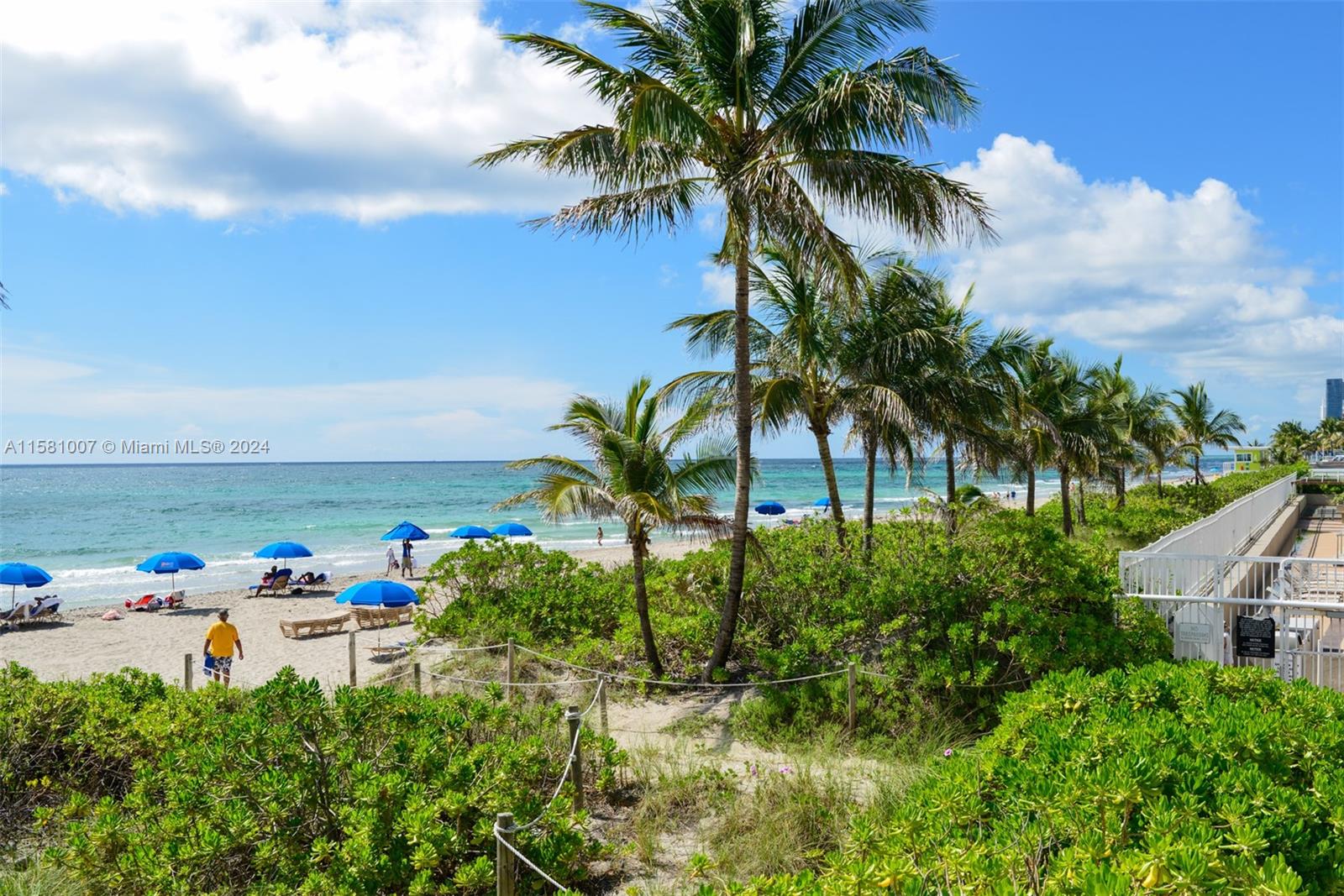 A sandy beach with palm trees, blue umbrellas, and a wooden walkway railing — 1950 S Ocean Dr # 21G, Hallandale Beach FL 33009, HALLANDALE BEACH, FL