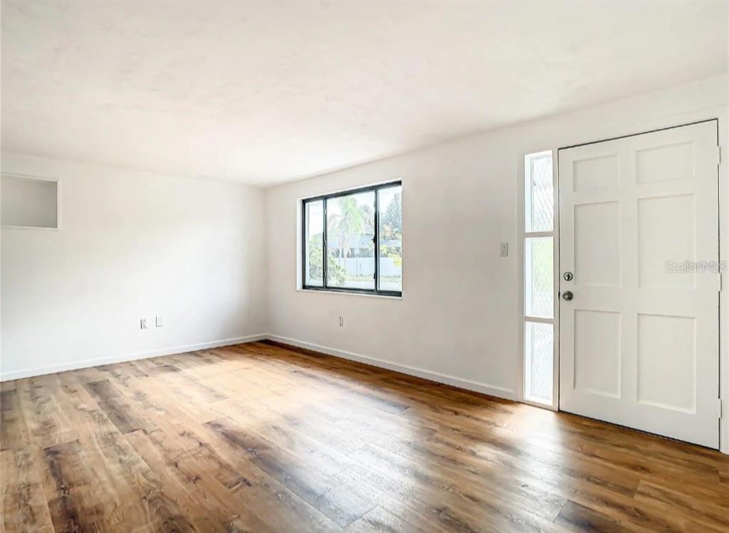 An empty room features white walls, wood-look flooring, a window, and a white front door — 4731 PHOENIX AVENUE, HOLIDAY, FL