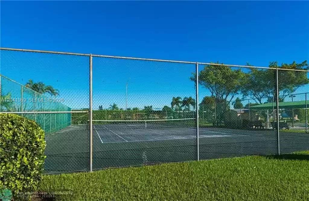 A fenced-in outdoor tennis court sits on a grassy lawn under a clear blue sky — 6870 Royal Palm Blvd # 111M, Margate FL 33063, MARGATE, FL