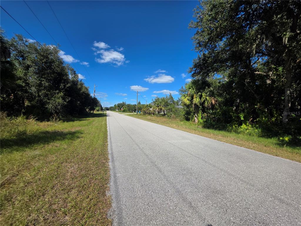 A paved road stretches into the distance, flanked by grassy shoulders and dense green trees — 414 LIDDY STREET, PORT CHARLOTTE, FL