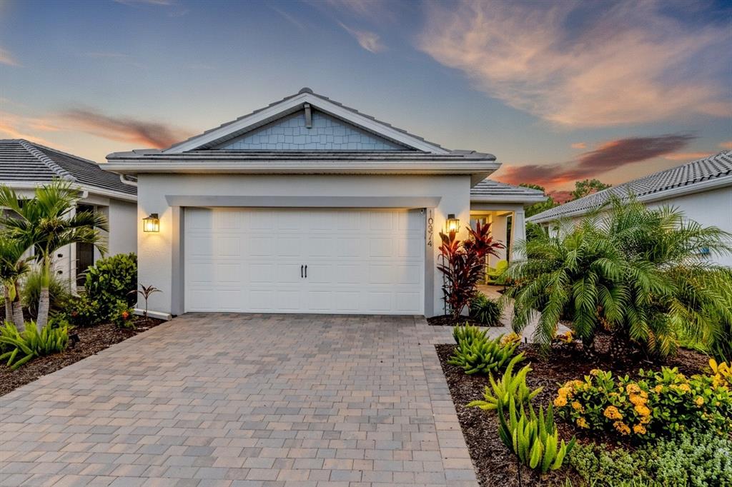 A single-story house with a white garage, paver driveway, and tropical landscaping at sunset — 10374 ABACO FALLS DRIVE, ENGLEWOOD, FL