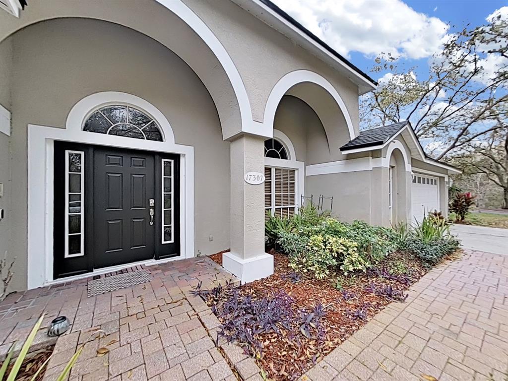 A beige house exterior features a dark front door, arched entryways, and a paver driveway — 17307 OAK LEDGE DRIVE, LUTZ, FL