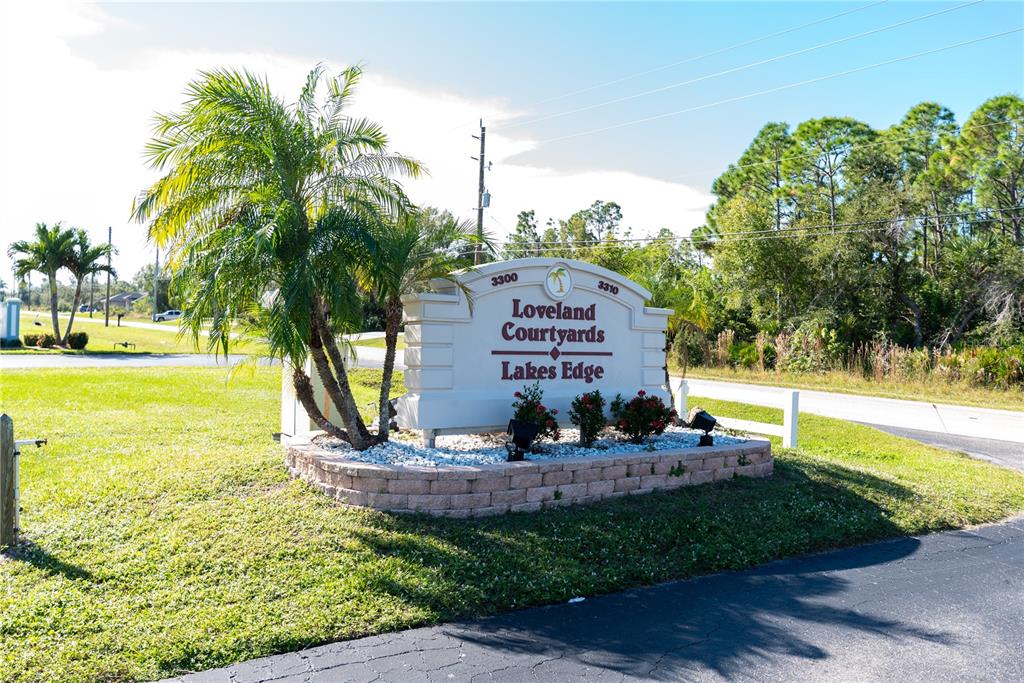 A sign for Loveland Courtyards Lakes Edge sits in a landscaped bed with palm trees — 3300 LOVELAND BOULEVARD UNIT 2103, PUNTA GORDA, FL
