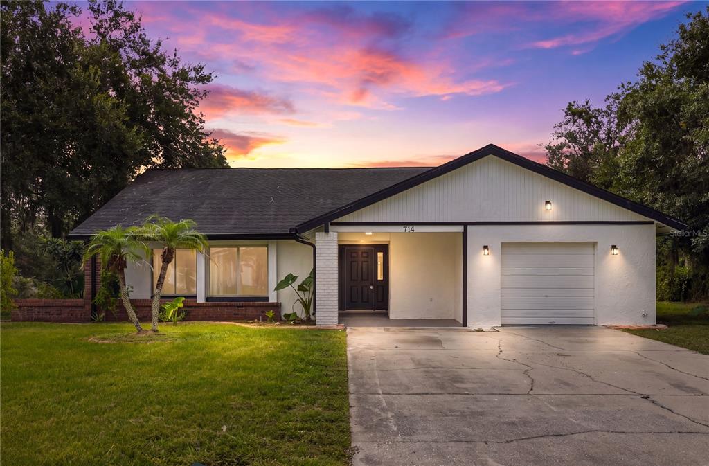 A single-story house with a white exterior, a dark roof, and a concrete driveway at sunset — 714 DIVOT LANE, POINCIANA, FL