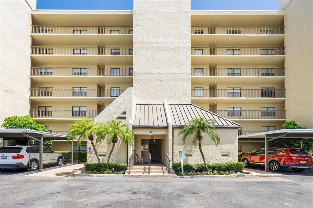 A multi-story apartment building with a central entrance, covered parking, and palm trees — 3300 COVE CAY DRIVE UNIT 7D, CLEARWATER, FL