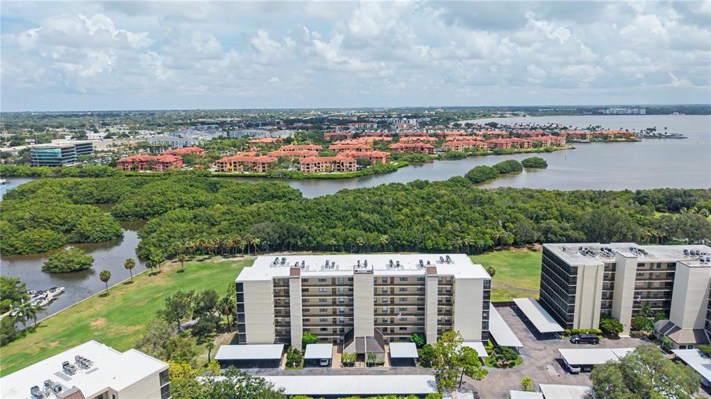 An aerial view shows multi-story apartment buildings surrounded by green space, mangroves, and a bay — 3300 COVE CAY DRIVE UNIT 7D, CLEARWATER, FL