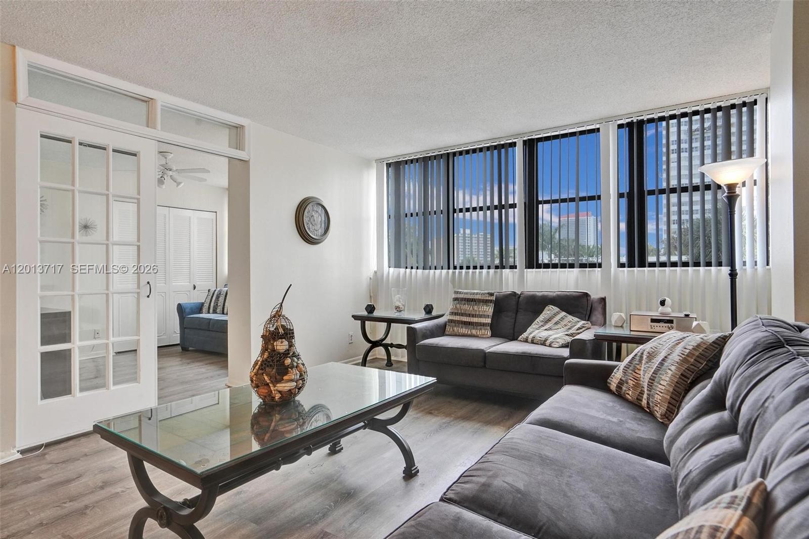 A living room features gray sofas, a glass coffee table, and large windows with blinds — 2017 S Ocean Dr # 205, Hallandale Beach FL 33009, HALLANDALE BEACH, FL
