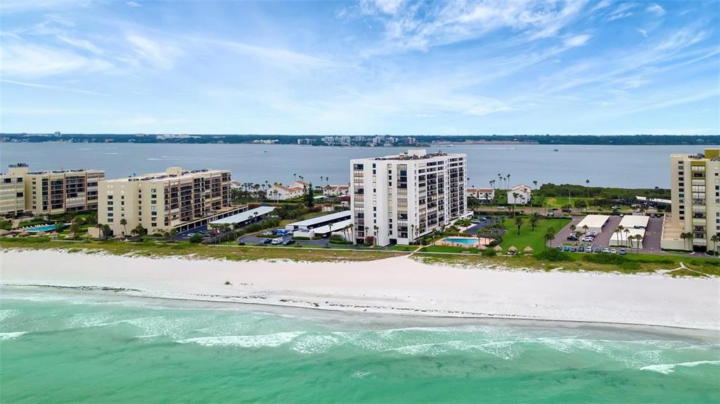 An aerial view shows beachfront apartment buildings with swimming pools and parking lots near water — 1460 GULF BOULEVARD UNIT 308, CLEARWATER BEACH, FL
