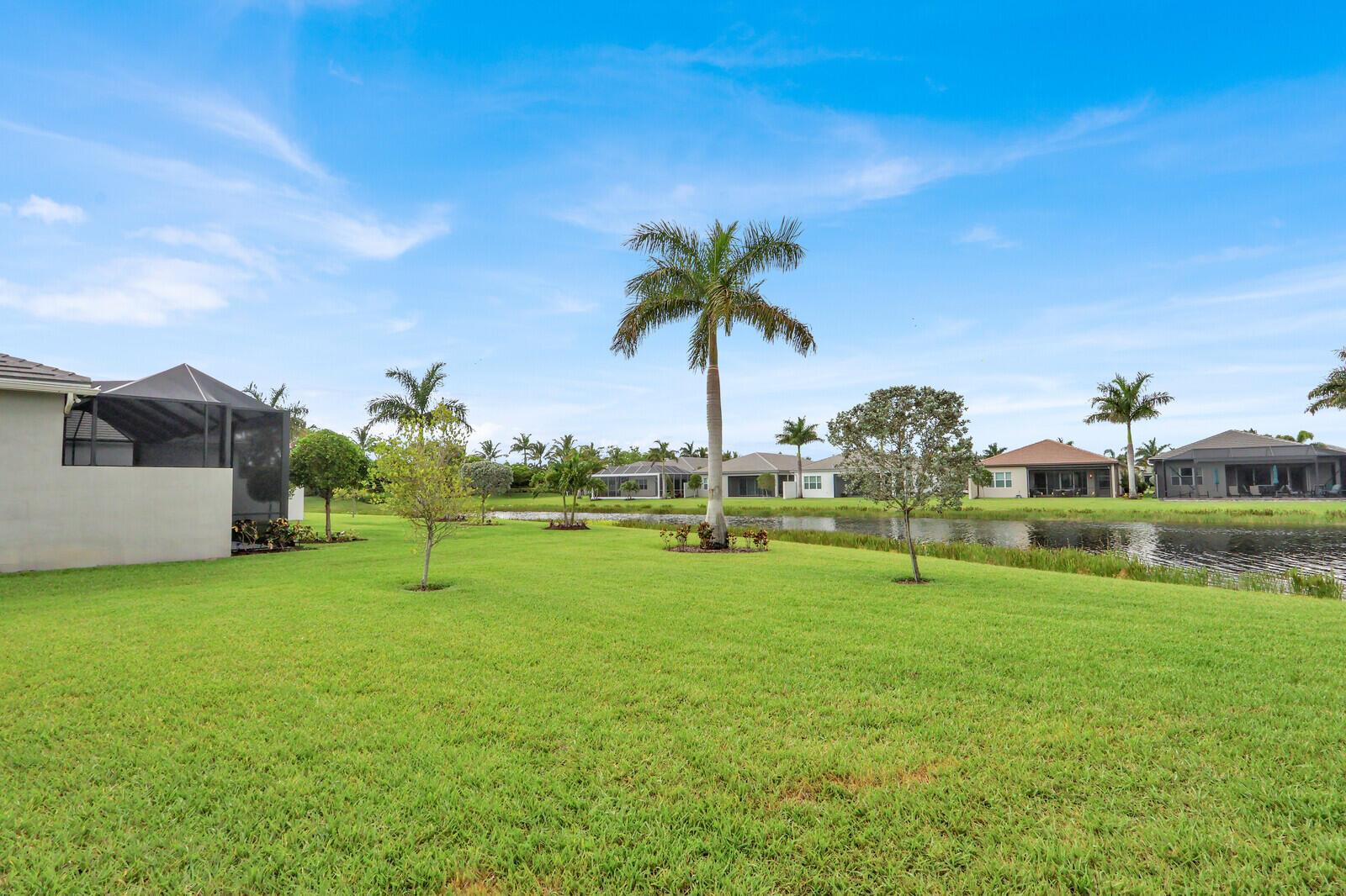 A grassy backyard features a palm tree, a small pond, and several houses in the distance — 9939 Great Barrier Trail, Boynton Beach FL 33473, BOYNTON BEACH, FL