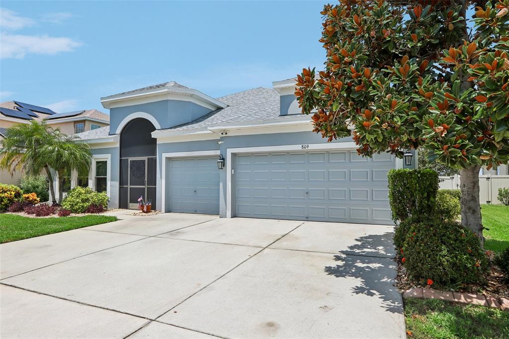 A light blue single-story house features a three-car garage and a large magnolia tree — 809 SEMINOLE SKY DRIVE, RUSKIN, FL