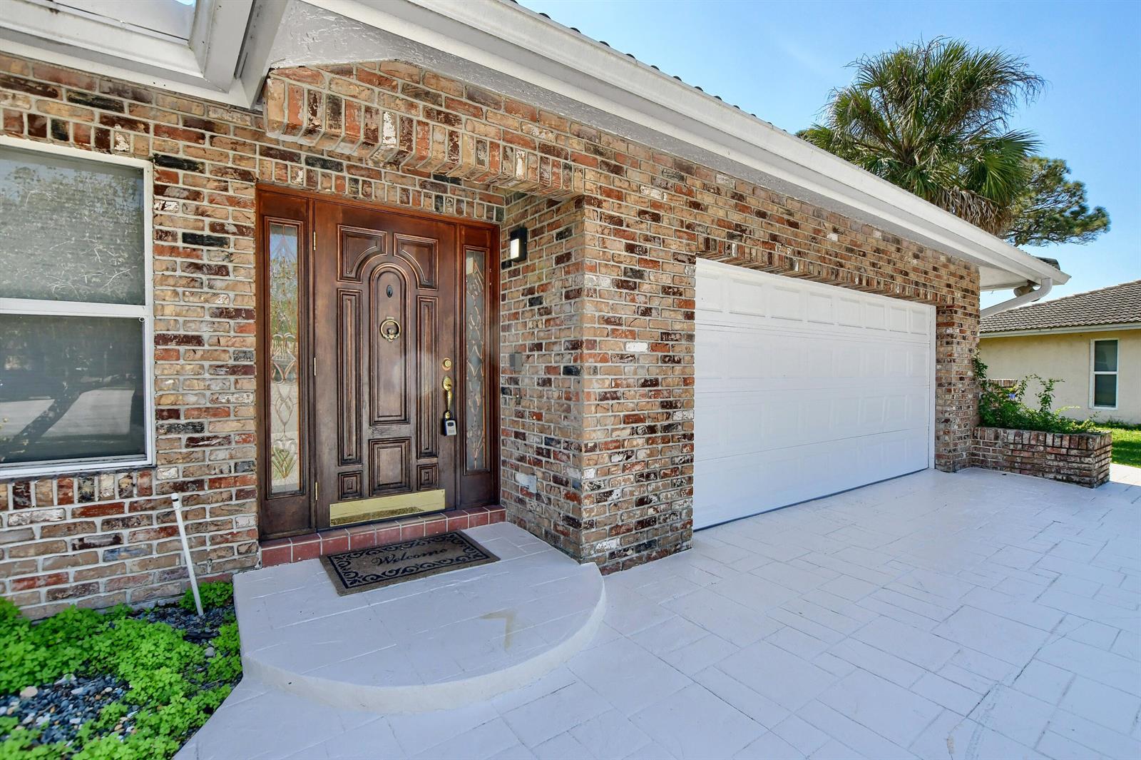 A brick-front house exterior features a wooden front door, a white garage door, and pavers — 2334 Timbercreek Circle NW, Boca Raton FL 33431, BOCA RATON, FL
