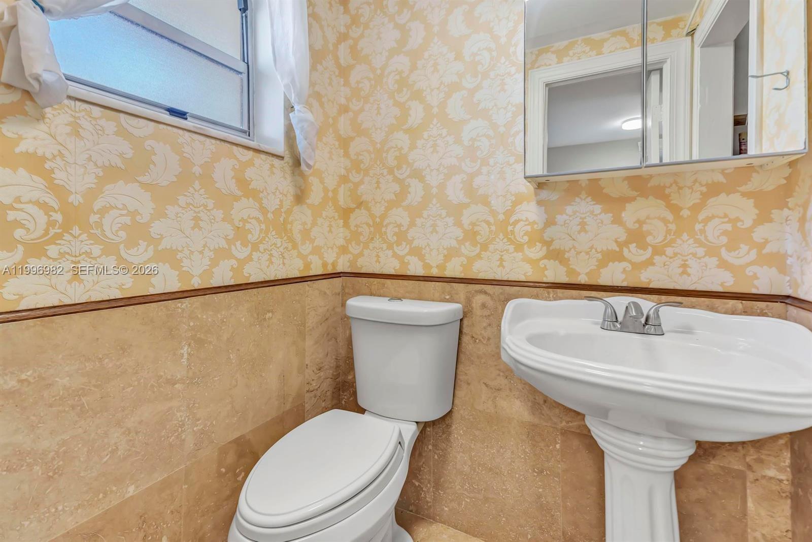 A bathroom features a pedestal sink, a toilet, beige tile wainscoting, and patterned wallpaper — 3199 S Ocean Dr # 406, Hallandale Beach FL 33009, HALLANDALE BEACH, FL