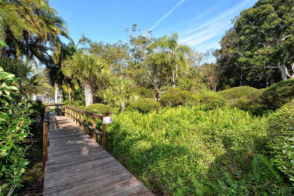 A wooden boardwalk winds through a lush, green garden with palm trees and dense foliage — 1608 BAYHOUSE POINT DRIVE UNIT GL304, SARASOTA, FL