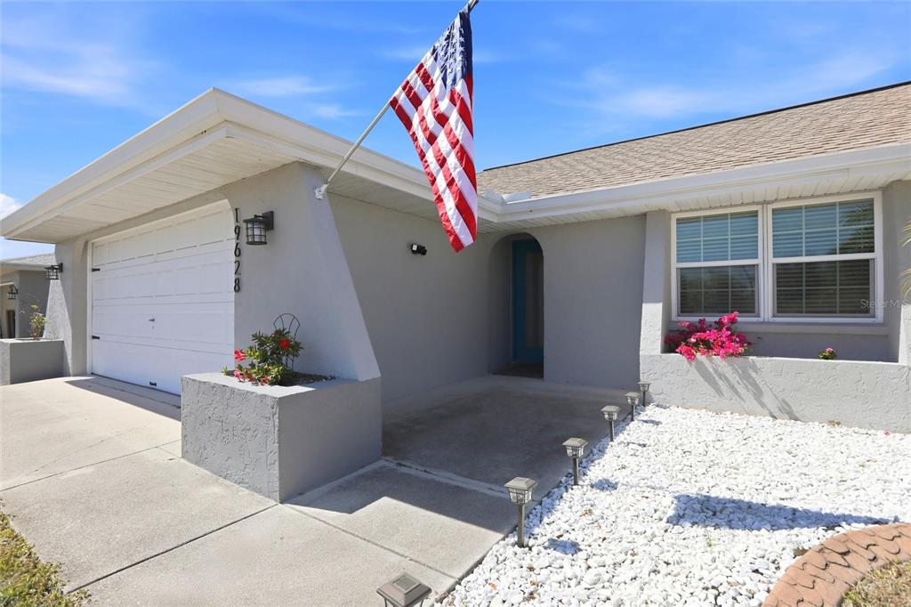 A gray house exterior features a white garage door, an American flag, and white gravel — 19628 MIDWAY BOULEVARD, PORT CHARLOTTE, FL