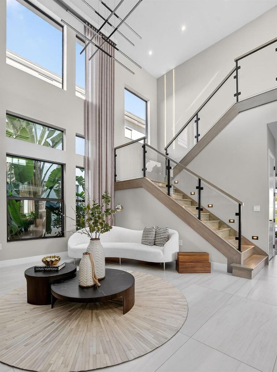 A modern foyer features a curved white sofa, a staircase with glass railings, and tall windows — 17080 Teton River Road, Boca Raton FL 33496, BOCA RATON, FL