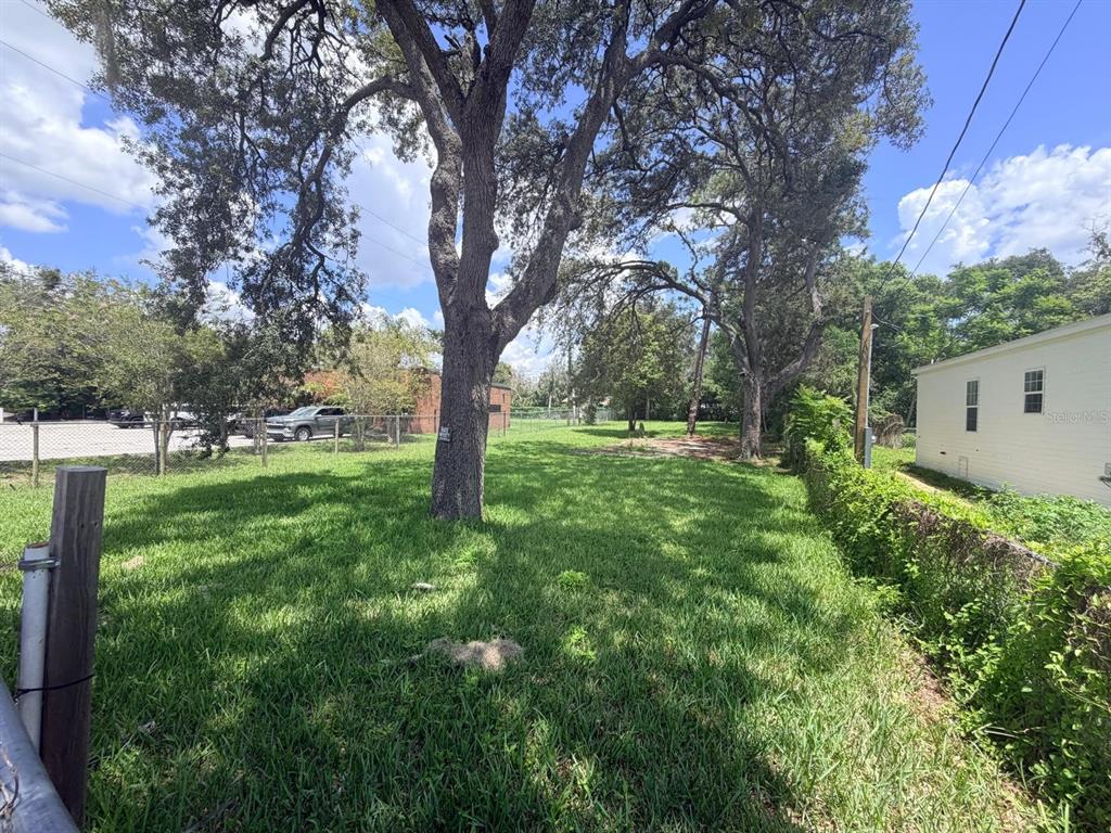 A grassy lot features large mature trees, a chain-link fence, and an adjacent white building — 1329 LAKE DRIVE, CASSELBERRY, FL