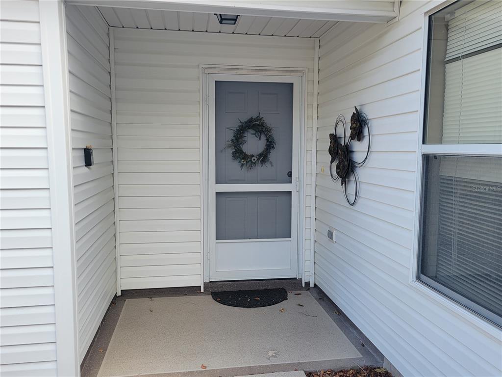A white front door with a screen door and wreath sits under a covered entryway — 1631 EDISTO LANE, THE VILLAGES, FL