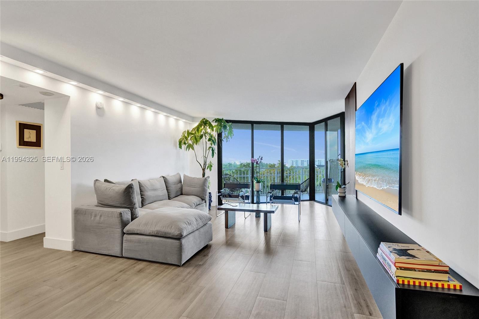A modern living room features a gray sectional sofa, wall-mounted television, and floor-to-ceiling windows — 1000 Quayside Ter # 707, Miami FL 33138, MIAMI, FL