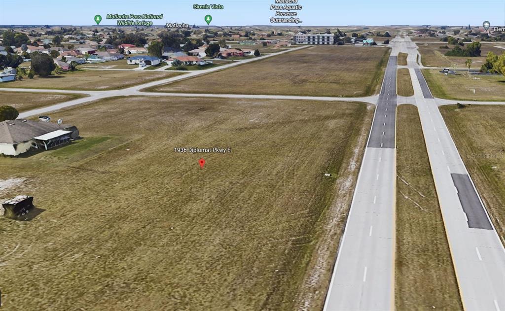 This aerial view shows a vacant, grassy lot adjacent to a multi-lane paved road — 1936 DIPLOMAT PARKWAY E, CAPE CORAL, FL