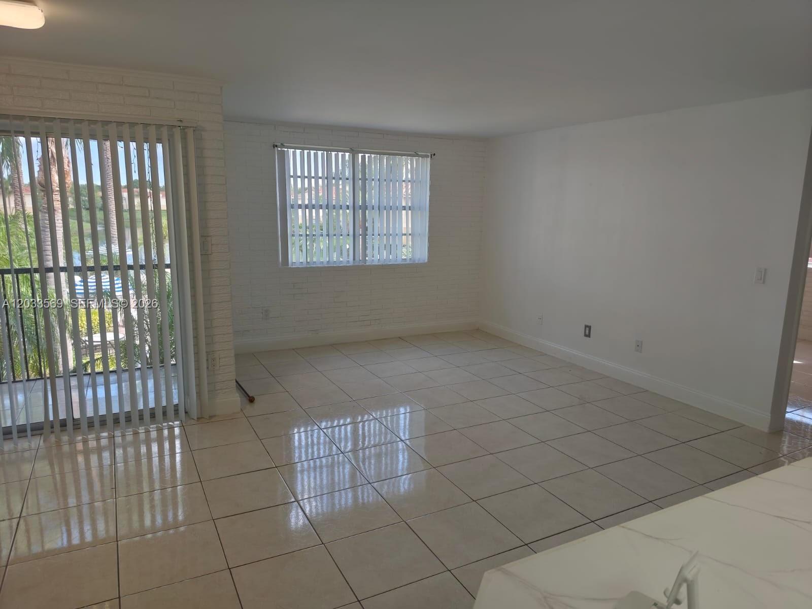 An empty living room features white brick walls, light tile flooring, and a sliding glass door — 1269 SW 46th Ave # 2408, Pompano Beach FL 33069, POMPANO BEACH, FL