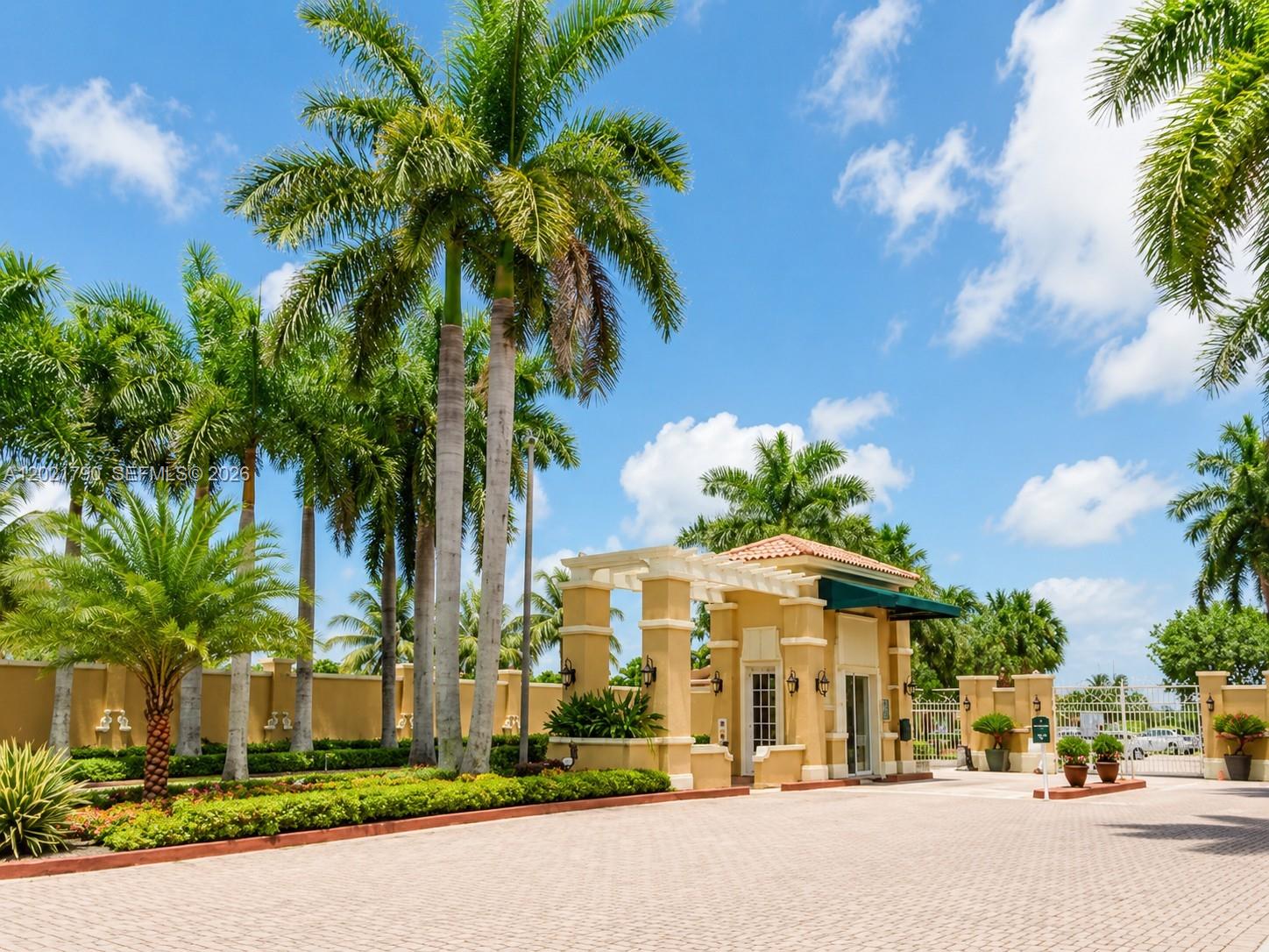 A gated entrance with palm trees, a paved driveway, and a tan guardhouse under blue skies — 6500 NW 114th Ave # 1021, Doral FL 33178, DORAL, FL