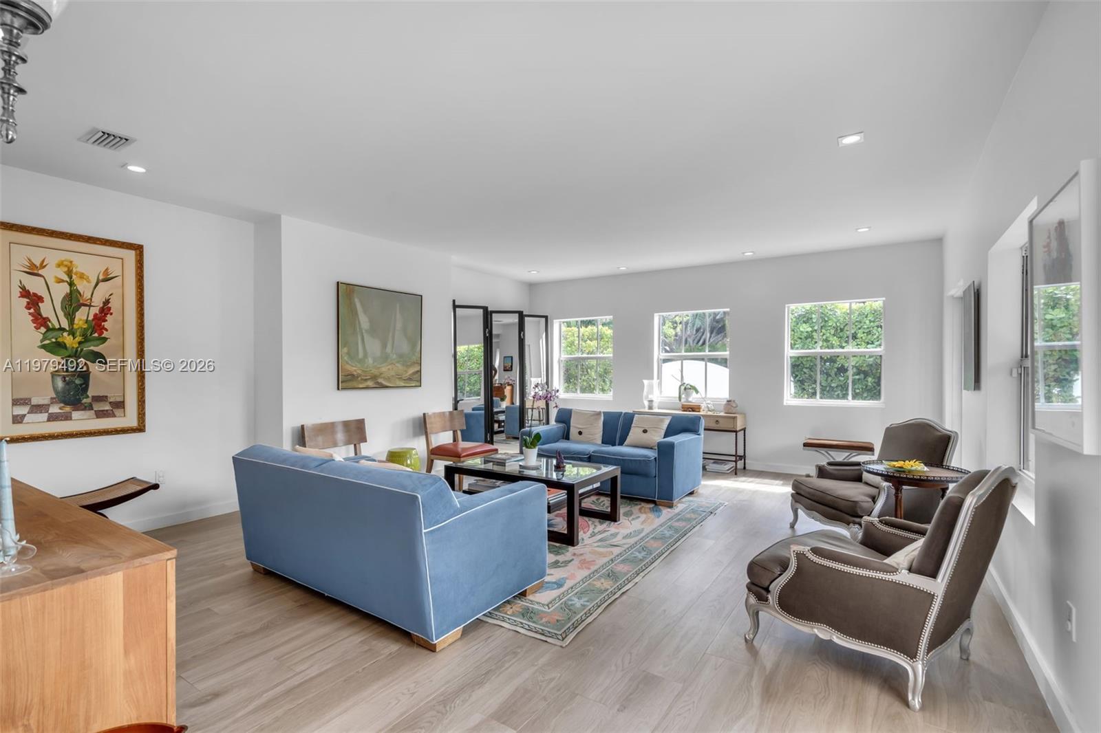 A bright living room features light wood flooring, blue sofas, and three windows — 707 W 54th St, Miami Beach FL 33140, MIAMI BEACH, FL