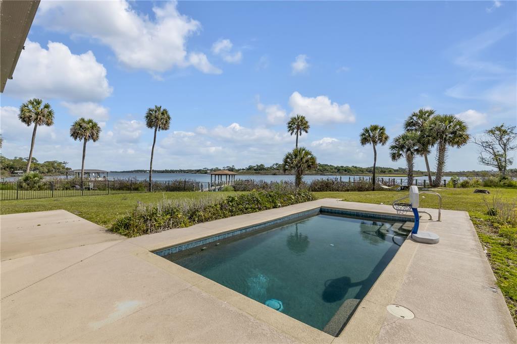 A rectangular swimming pool sits on a concrete patio overlooking a lake with palm trees — 2185 TURNBULL BAY ROAD, NEW SMYRNA BEACH, FL