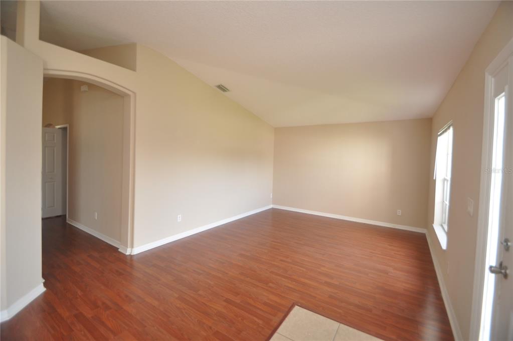 An empty living room features vaulted ceilings, beige walls, and dark wood-look laminate flooring — 37 LOUISBURG LANE, PALM COAST, FL