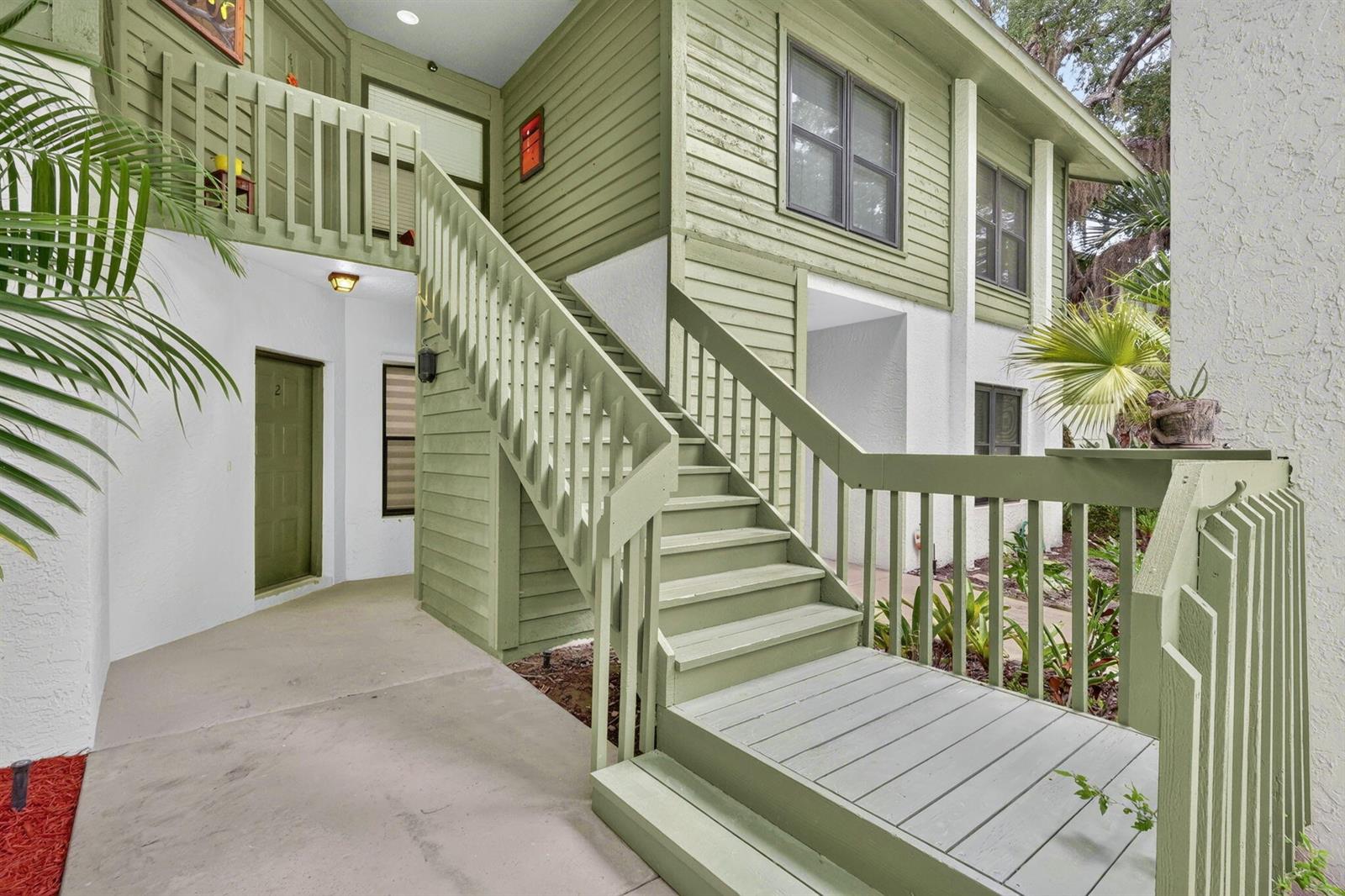 An exterior view of a two-story building with green siding, a wooden staircase, and landscaping — 5250 SE Seascape Way # 102, Stuart FL 34997, STUART, FL