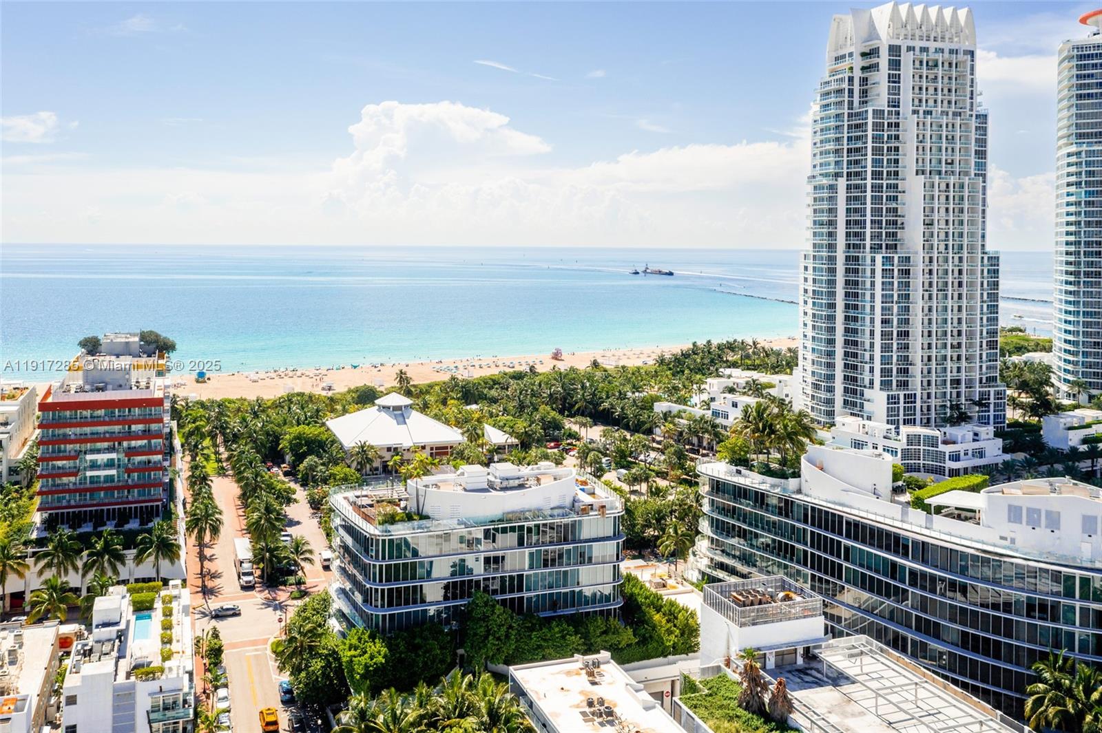 An aerial view shows a coastal city with high-rise buildings, lush trees, and a sandy beach — 1 Collins Ave # 207, Miami Beach FL 33139, MIAMI BEACH, FL