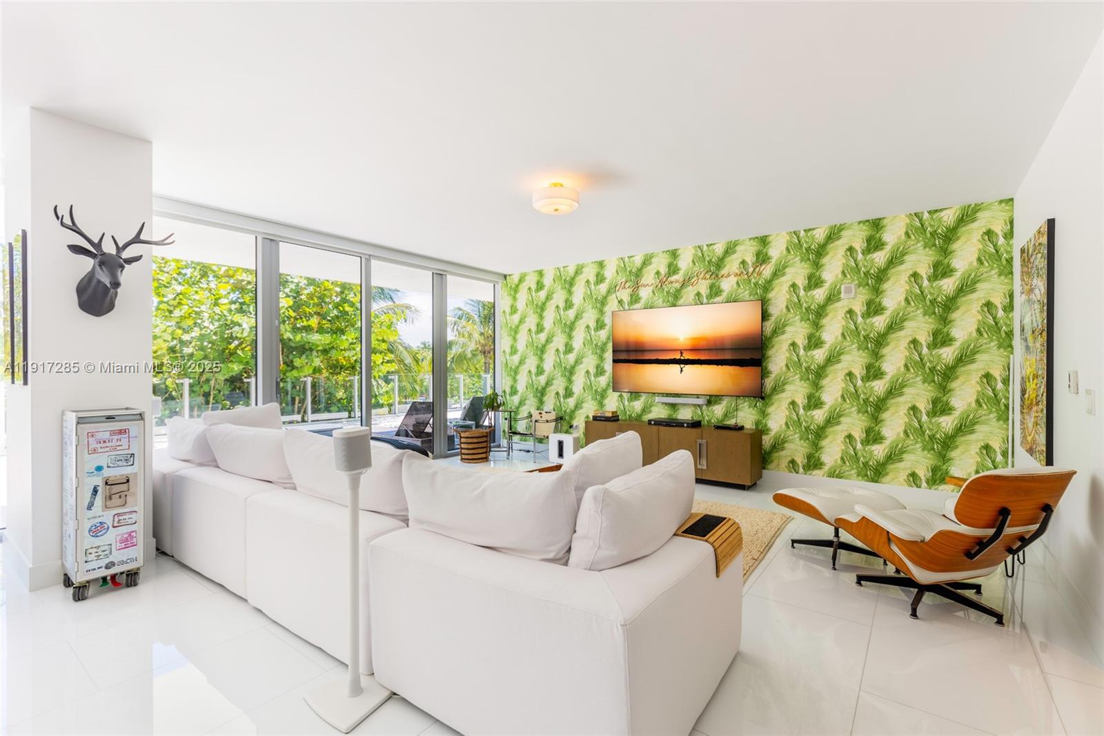 A living room features white sofas, a palm-leaf wallpaper accent wall, and floor-to-ceiling glass windows — 1 Collins Ave # 207, Miami Beach FL 33139, MIAMI BEACH, FL