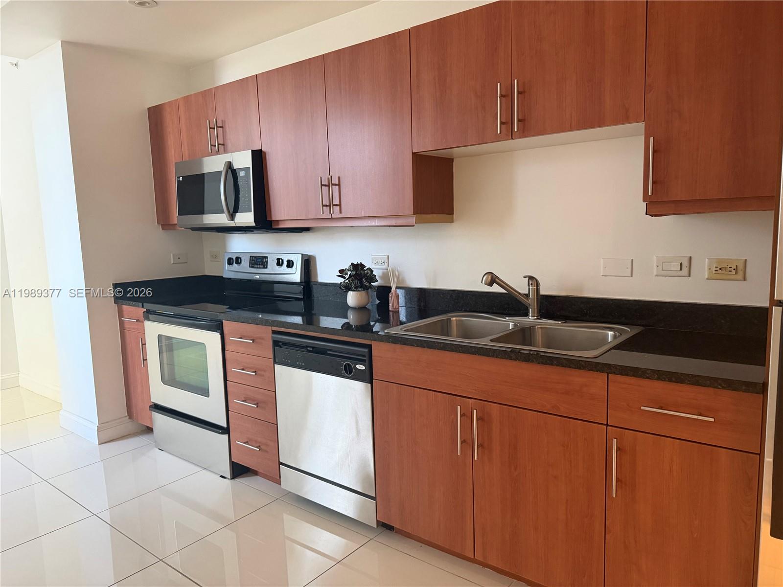 A kitchen features wood cabinets, stainless steel appliances, black countertops, and white tile flooring — 111 E Flagler St # 606, Miami FL 33131, MIAMI, FL