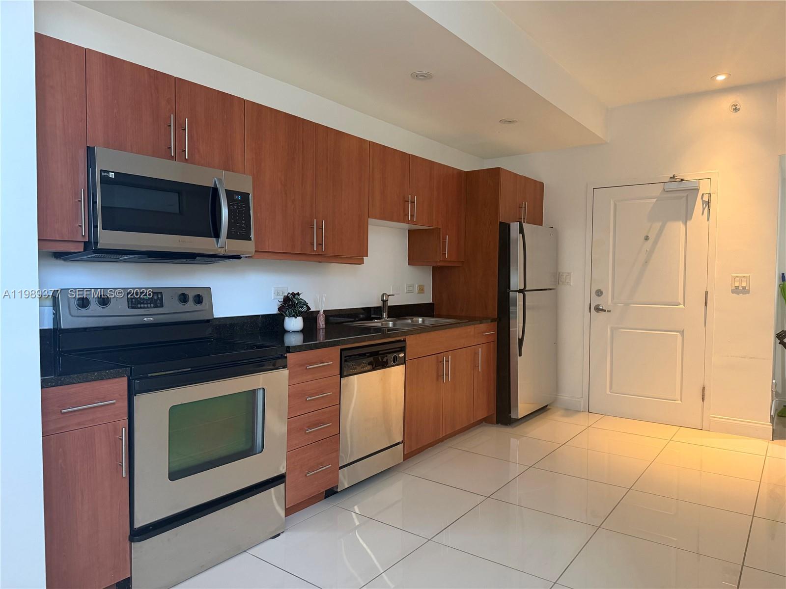 A kitchen features wood-toned cabinetry, stainless steel appliances, black countertops, and white tile flooring — 111 E Flagler St # 606, Miami FL 33131, MIAMI, FL