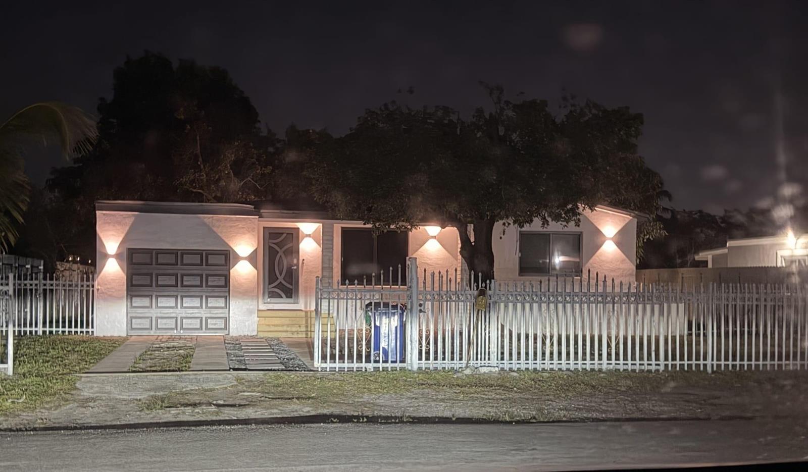 A single-story house with a white exterior, a garage, and a metal fence at night — 1025 NW 148th Street, Miami FL 33168, MIAMI, FL