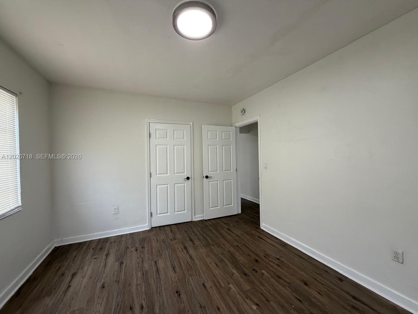 An empty bedroom features white walls, dark wood-look flooring, two white doors, and a ceiling light — MIAMI, FL