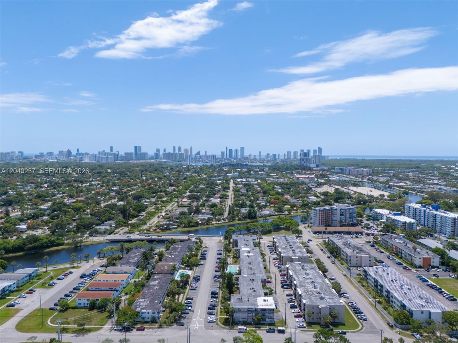 An aerial view shows residential apartment complexes and parking lots near a canal and city skyline — 1450 NE 170th St # 115, North Miami Beach FL 33162, NORTH MIAMI BEACH, FL