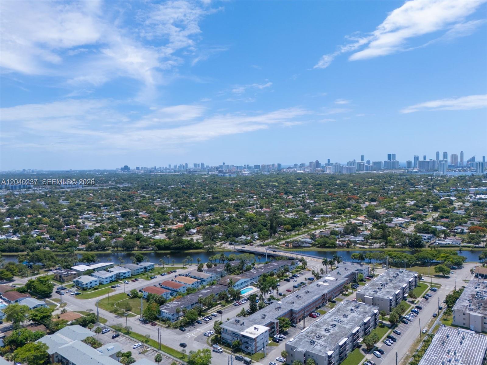 An aerial view shows a residential neighborhood with apartment complexes, a canal, and a distant skyline — 1450 NE 170th St # 115, North Miami Beach FL 33162, NORTH MIAMI BEACH, FL