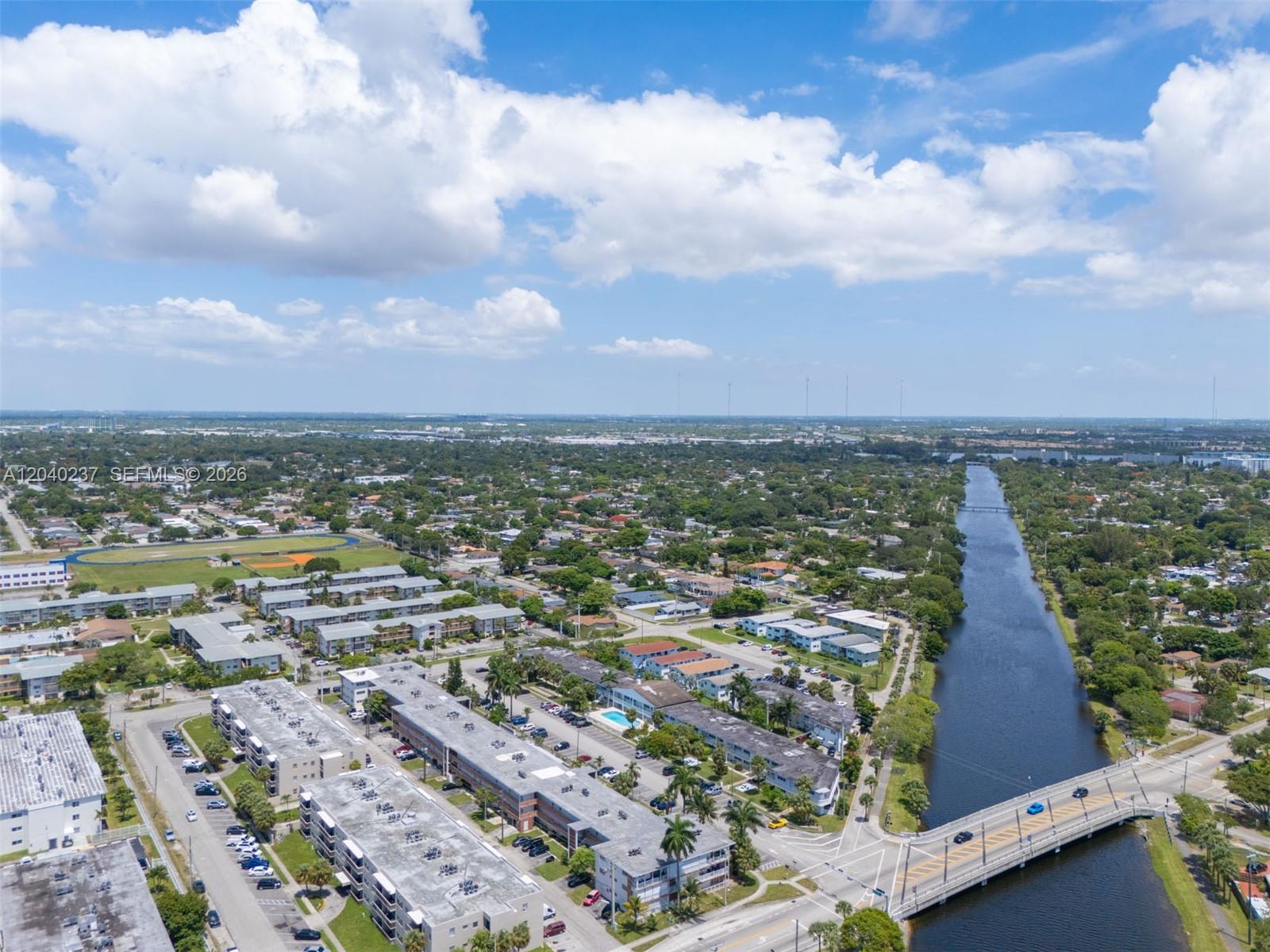 An aerial view shows residential apartment buildings, a canal, and a bridge under a cloudy sky — 1450 NE 170th St # 115, North Miami Beach FL 33162, NORTH MIAMI BEACH, FL