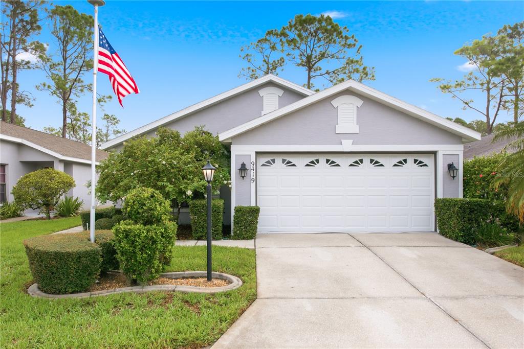 A gray single-story house with a two-car garage, a flag pole, and manicured landscaping — 9419 GREEN NEEDLE DRIVE, NEW PORT RICHEY, FL