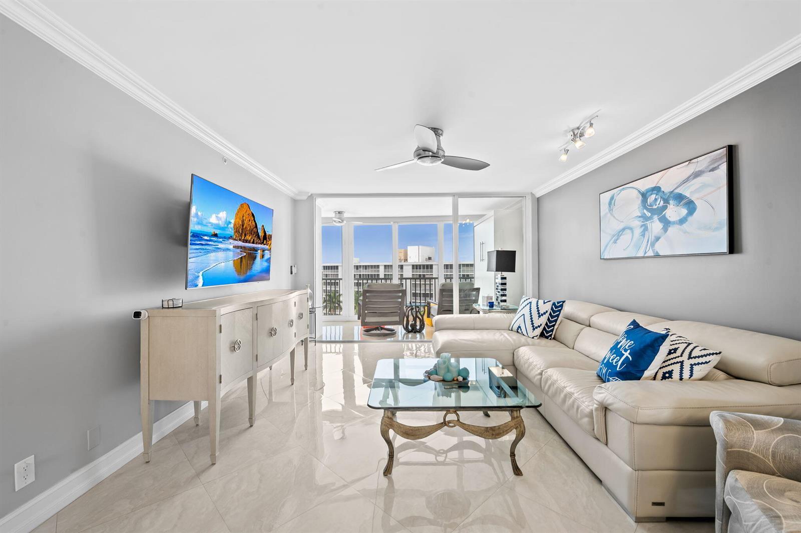 A living room features a white sectional sofa, glass coffee table, and a wall-mounted television — 3300 S Ocean Boulevard # 1020, Highland Beach FL 33487, HIGHLAND BEACH, FL