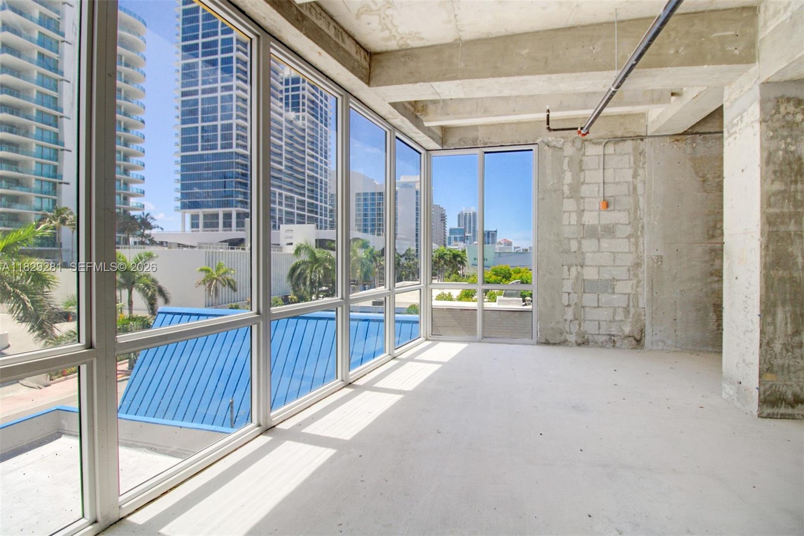 An unfinished room features concrete walls, exposed ceiling pipes, and floor-to-ceiling windows overlooking a city — 6936 Collins Ave, Miami Beach FL 33141, MIAMI BEACH, FL