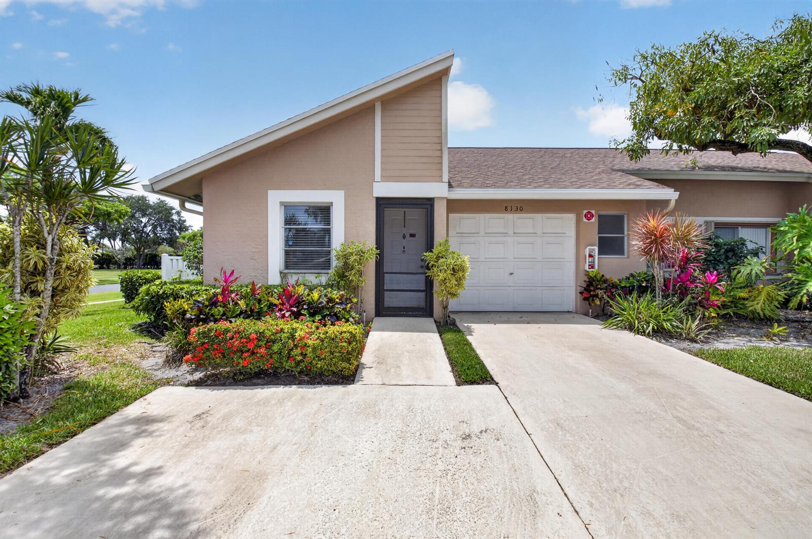 A single-story house with a beige stucco exterior, a white garage door, and lush landscaping — 8130 Springtree Road # A, Boca Raton FL 33496, BOCA RATON, FL