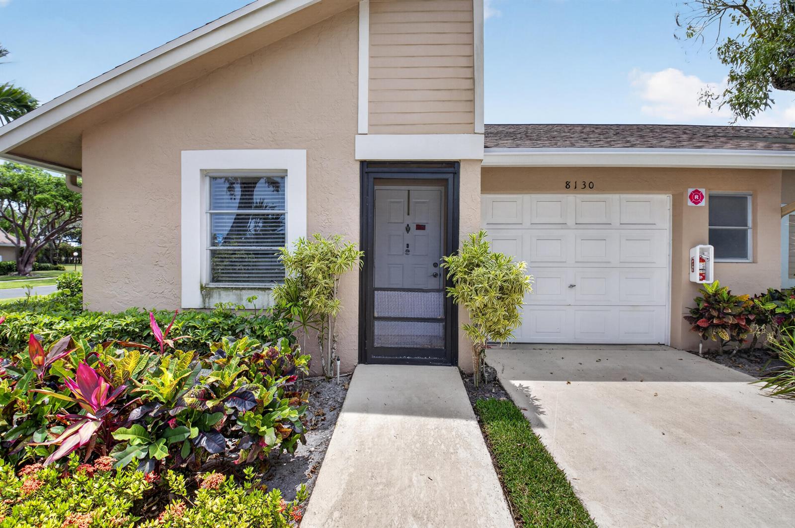 The exterior of a single-story home features a tan stucco facade, a garage, and landscaping — 8130 Springtree Road # A, Boca Raton FL 33496, BOCA RATON, FL