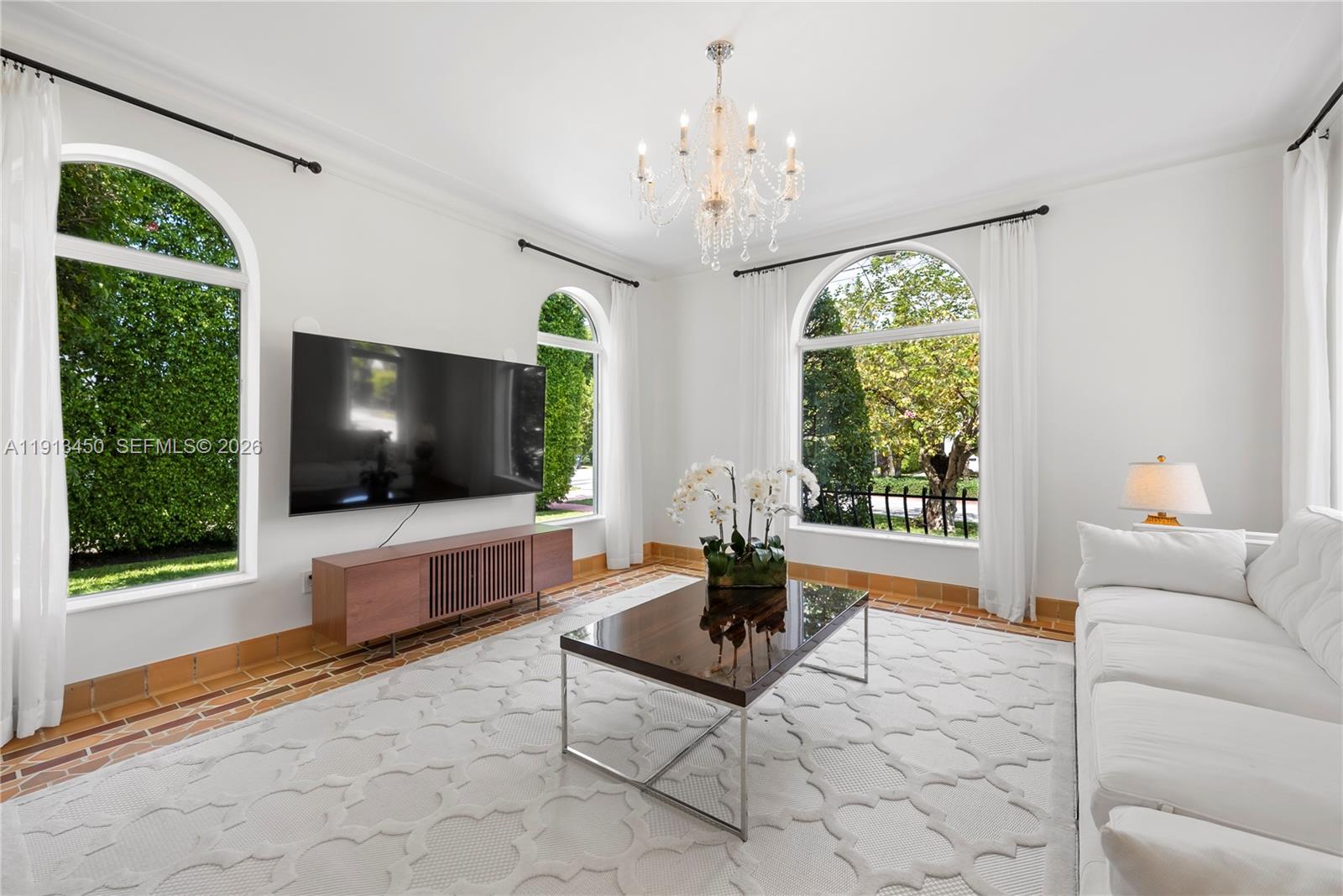 A bright living room features a large television, white sofa, chandelier, and arched windows — 5800 La Gorce Dr, Miami Beach FL 33140, MIAMI BEACH, FL