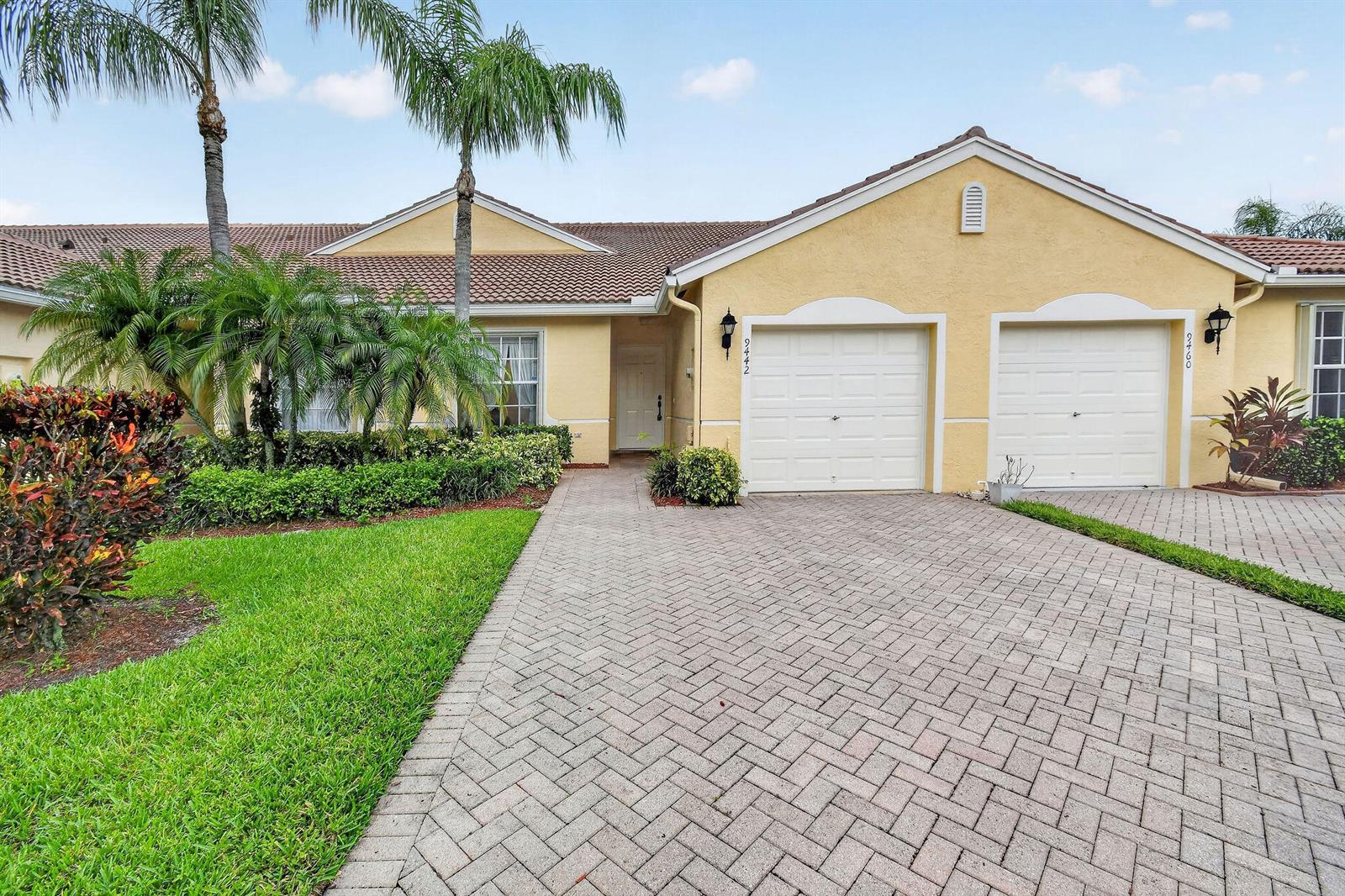 A yellow stucco house features a paver driveway, two garage doors, and palm trees — 9442 Bridgeport Drive, West Palm Beach FL 33411, WEST PALM BEACH, FL