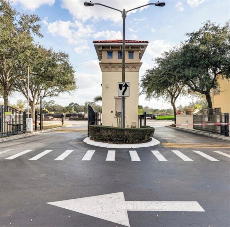 A gated entrance features a central guardhouse, paved roads, and mature trees under a sky — 2704 CLUB CORTILE CIRCLE, KISSIMMEE, FL