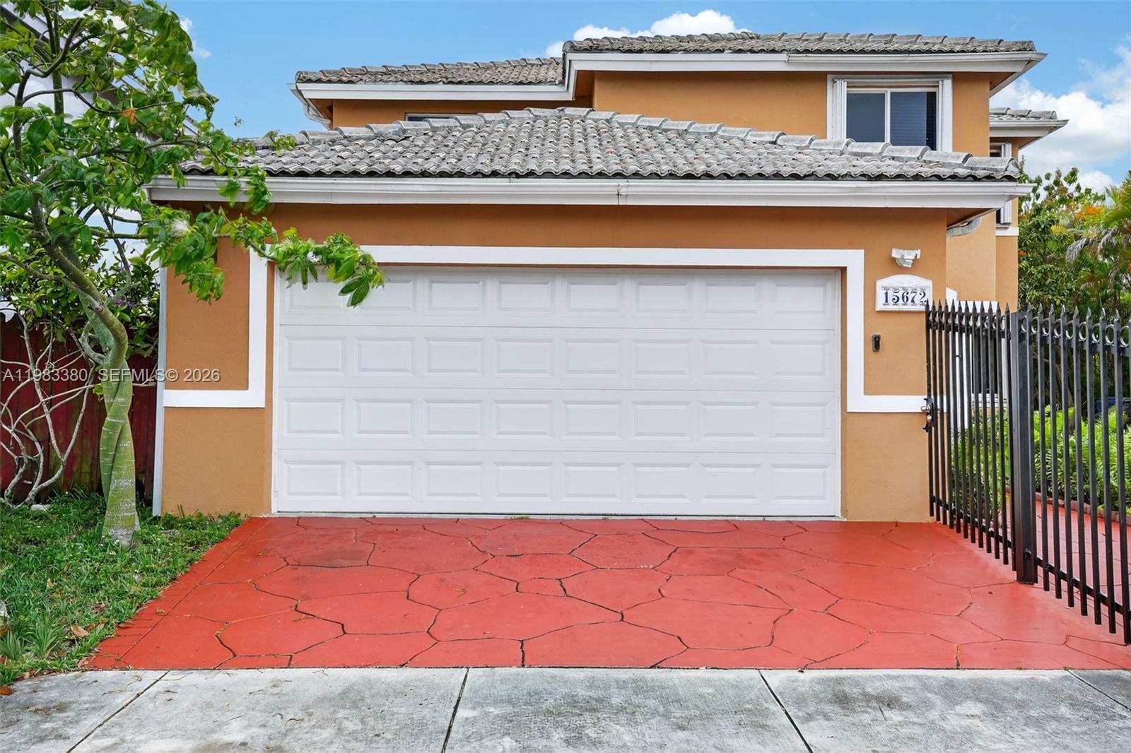 A two-story house exterior features a white garage door, red paved driveway, and metal fence — 15672 SW 91st Ln, Miami FL 33196, MIAMI, FL