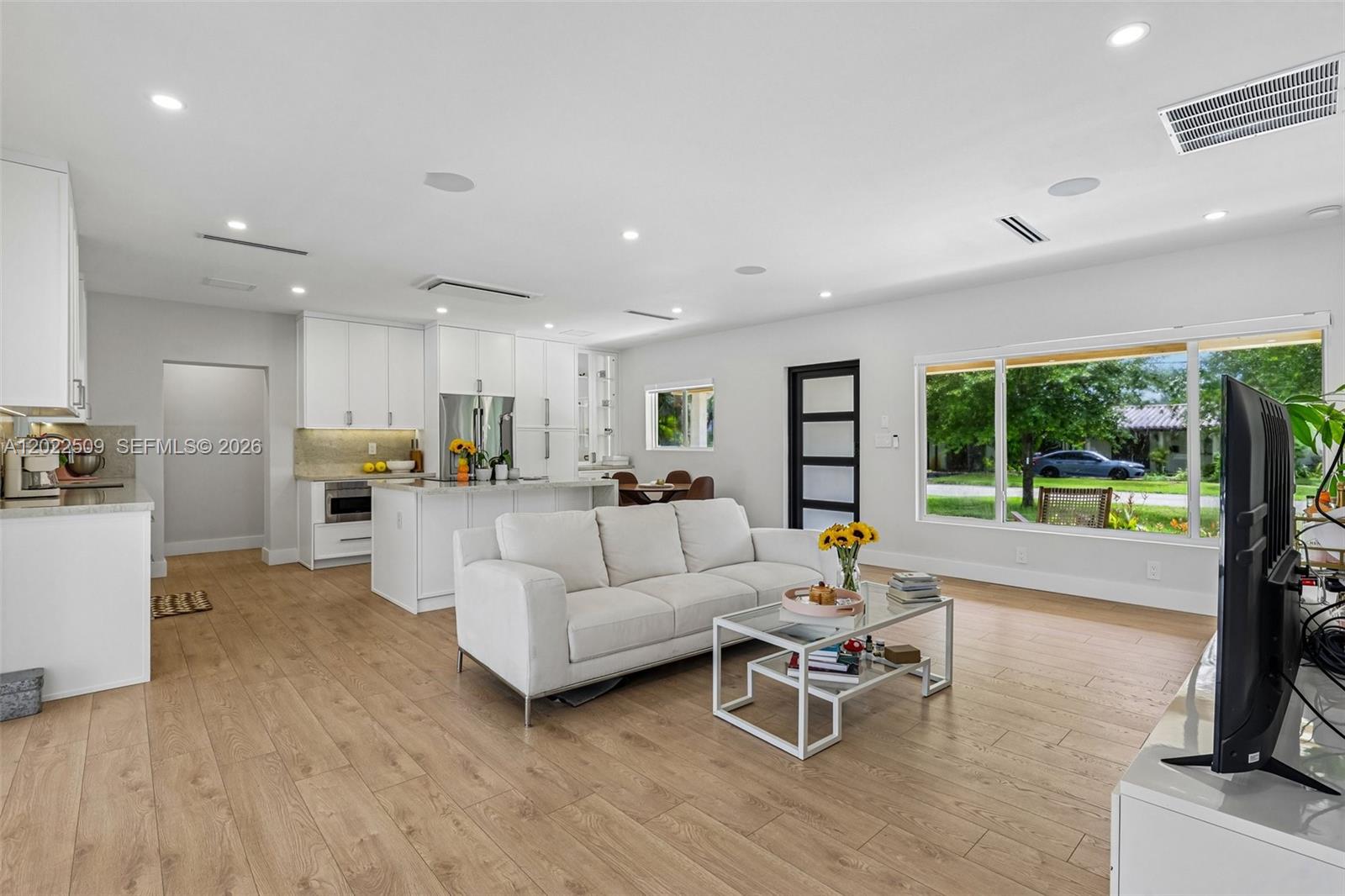 An open-concept living room and kitchen feature white cabinetry, light wood flooring, and large windows — 5620 SW 63rd Ct, South Miami FL 33143, SOUTH MIAMI, FL