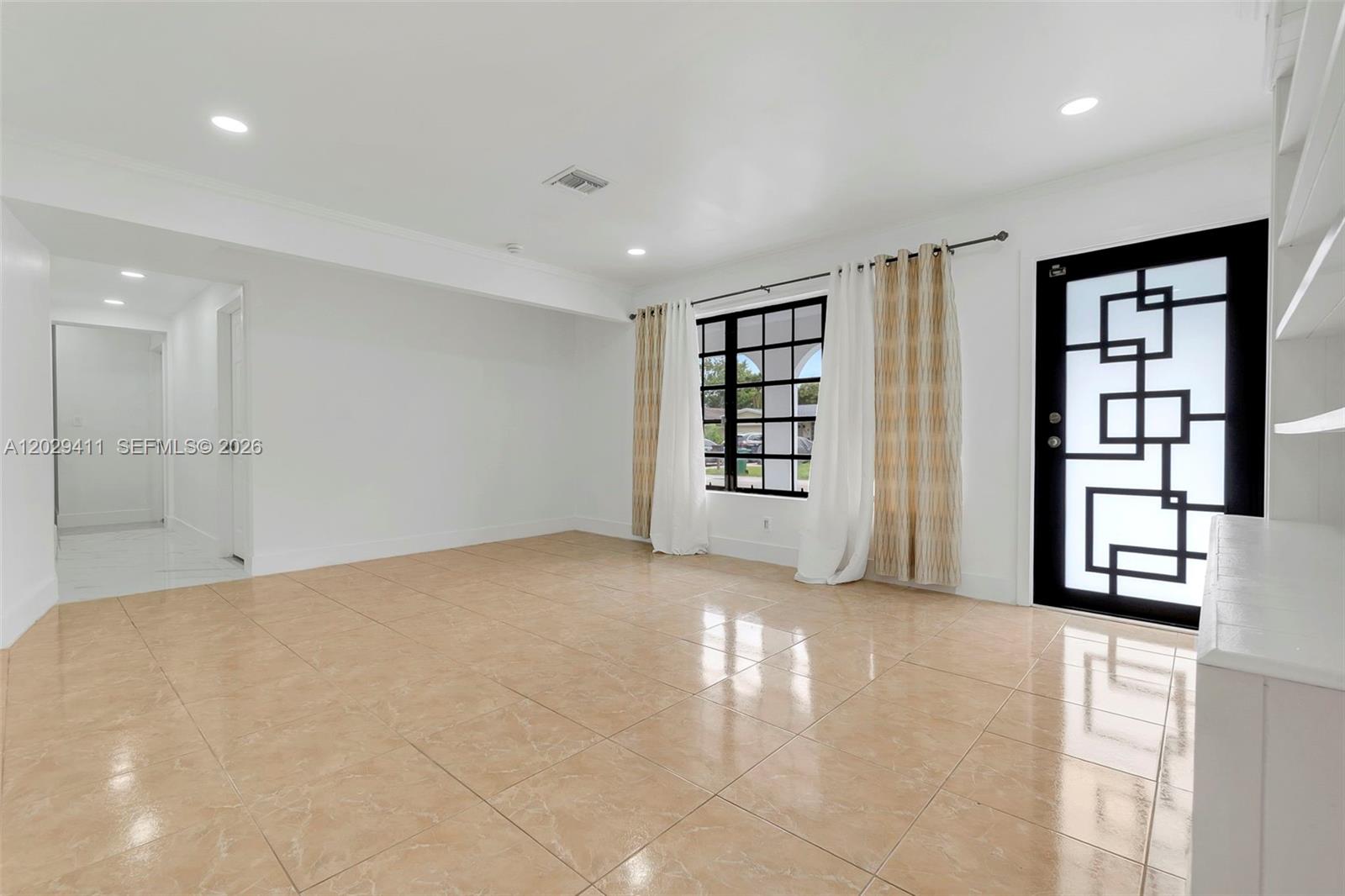 An empty room features beige tile flooring, white walls, a black geometric door, and curtains — 6101 SW 24th St # 1, Miramar FL 33023, MIRAMAR, FL
