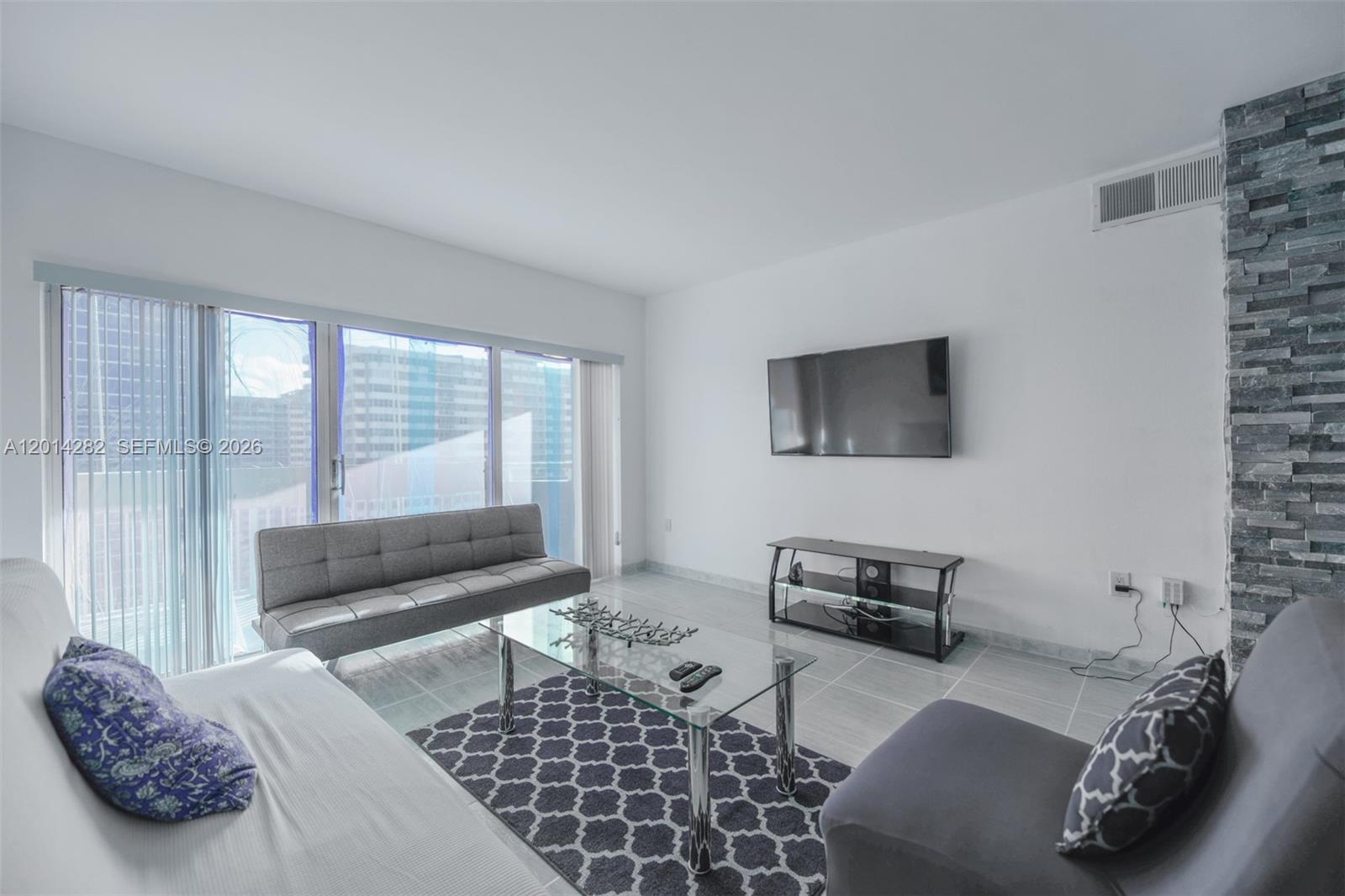 A living room features a glass coffee table, gray seating, a wall-mounted television, and tile flooring — 1950 S Ocean Dr # 17N, Hallandale Beach FL 33009, HALLANDALE BEACH, FL